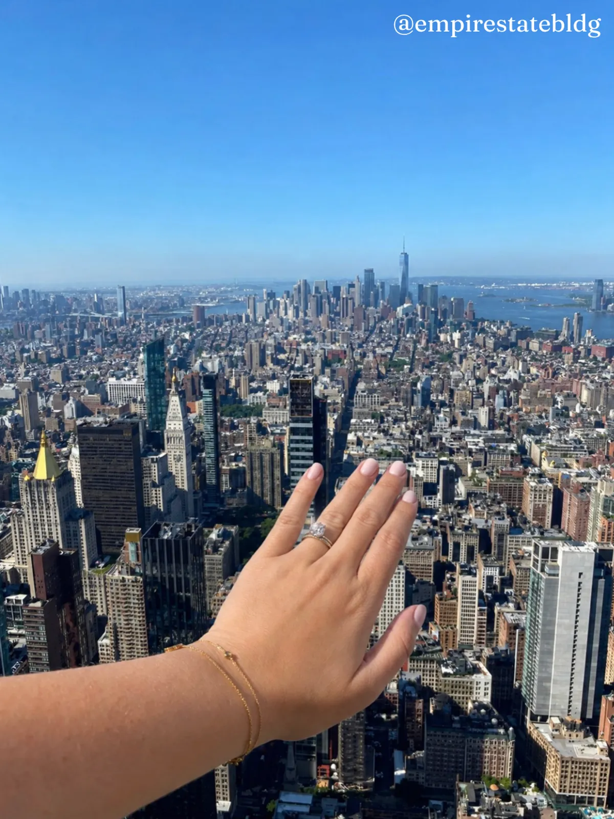 A person's outstretched hand with an engagement ring and gold bracelet reaches out from the camera in the foreground, showing off the ring against a sweeping aerial view of the New York City skyline, including One World Trade Center in the distance, with the "@empirestatebldg" handle in the top corner.