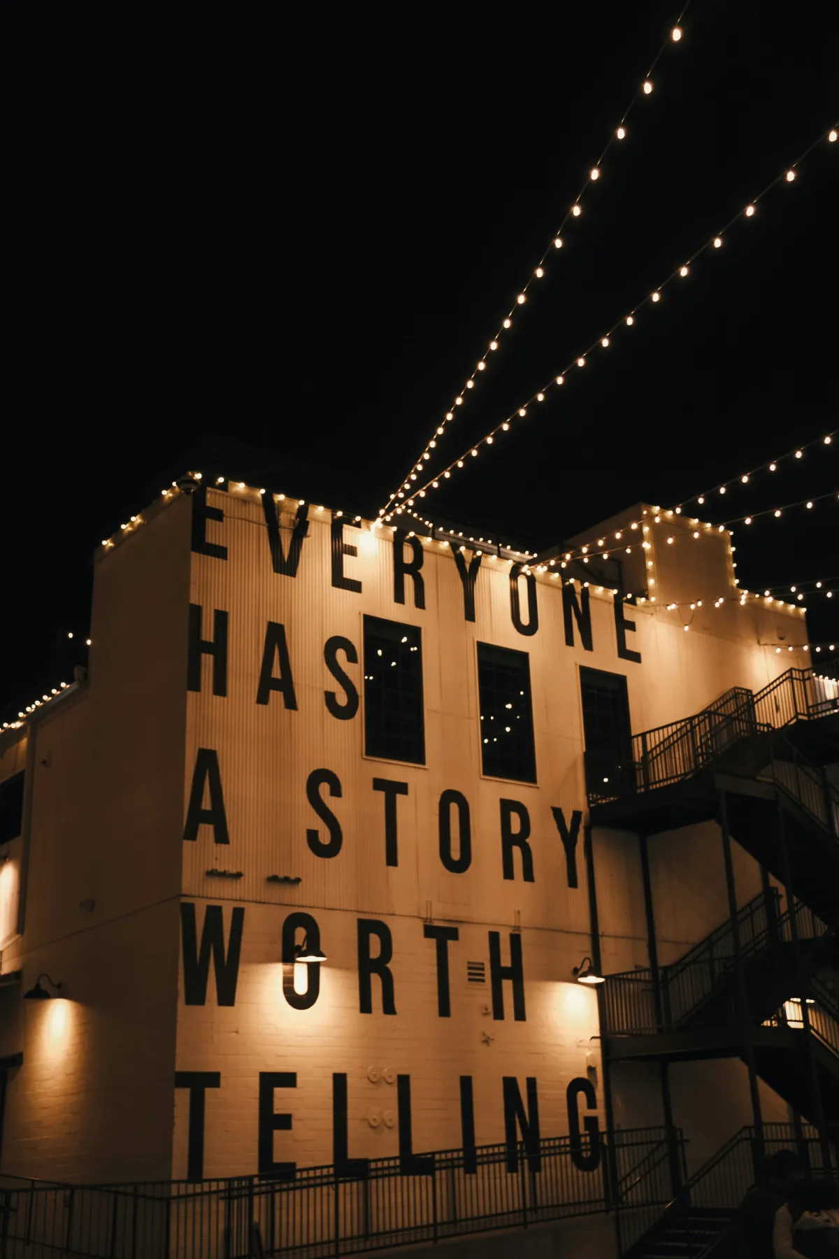 Illuminated building at night with string lights and the message “Everyone has a story worth telling” painted across its exterior.